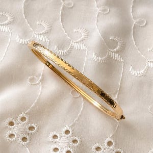 May include: A gold bangle bracelet with a textured, hammered design. The bracelet is set against a white, embroidered fabric background. The bracelet is a classic piece of jewellery.