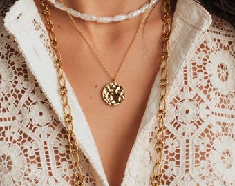 May include: A layered necklace set featuring a short strand of white freshwater pearls, a delicate gold chain with a round textured coin pendant, and a long gold chain with large oval links, worn over a white lace shirt with floral patterns.