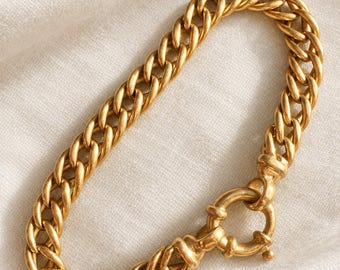 May include: A gold-toned chain bracelet featuring thick, interlocking oval links with a matte finish. The bracelet is secured by a large, circular spring-ring clasp that connects to the main chain, creating a continuous, uniform loop design.