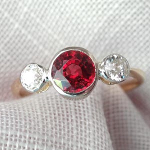 May include: A gold ring featuring a large, round, vibrant red gemstone flanked by two smaller, clear, round gemstones. The ring is set against a soft, white fabric background, highlighting the sparkle of the gems.