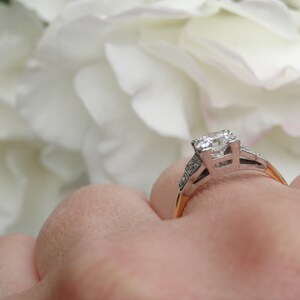 May include: A silver engagement ring with a large, clear diamond center stone and smaller diamonds on the band. The ring is on a finger and the background is blurred white flowers.