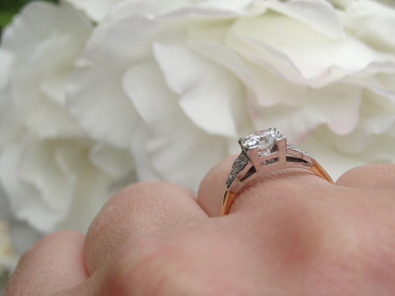 May include: A silver engagement ring with a large, clear diamond center stone and smaller diamonds on the band. The ring is on a finger and the background is blurred white flowers.