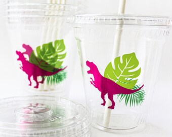 May include: Clear plastic cups with lids and straws. Each cup features a pink dinosaur silhouette with green tropical leaves. A stack of lids and a bundle of straws are also visible.