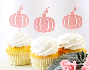 May include: Three cupcakes with pink glitter pumpkin toppers. The cupcakes are frosted with white frosting and have a yellow cake base.