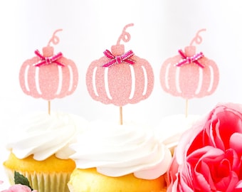 May include: Three cupcakes with white frosting and pink glitter pumpkin toppers with pink bows. A pink rose is in the foreground. The cupcakes have yellow cake and are in white paper liners.