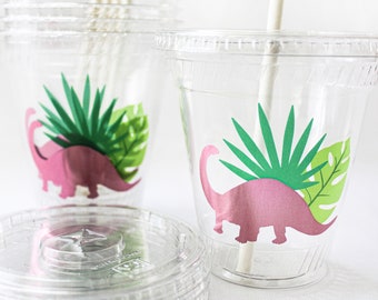 May include: Clear plastic cups with lids and straws. Each cup features a pink dinosaur illustration with green and dark green tropical leaves. Perfect for a dinosaur-themed party or event.