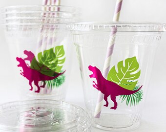 May include: Clear plastic cups with lids, decorated with a magenta dinosaur and green tropical leaves. The cups have striped straws. A stack of lids is in the foreground.