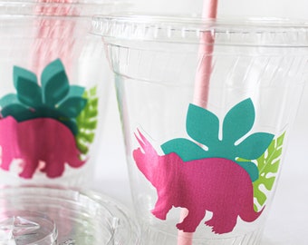 May include: Clear plastic cups with lids and pink paper straws. Each cup features a pink dinosaur silhouette with teal and green leaf accents. The cups are designed for beverages and are suitable for a party or event.