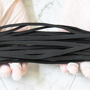 May include: A pile of black elastic cord, held by gloved hands. The cord is tightly woven and appears to be made of a stretchy material. The background is a light marble pattern.