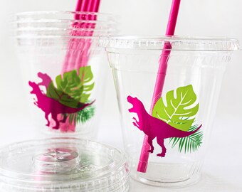 May include: Clear plastic cups with lids and pink straws. Each cup features a magenta dinosaur and green tropical leaf design. The cups are stacked, with some lids visible, suggesting a party or event theme.