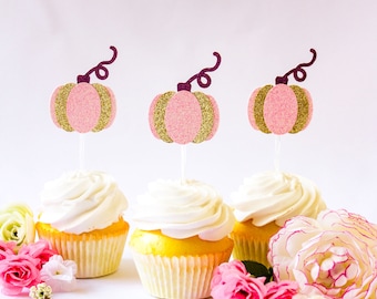 May include: Three cupcakes decorated with pink and gold glitter pumpkin toppers. The cupcakes have white frosting and are on a white surface.