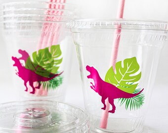 May include: Clear plastic cups with pink dinosaur and green leaf designs. Pink paper straws are in the cups. A stack of clear plastic lids is in the foreground. The cups are suitable for a dinosaur-themed party.