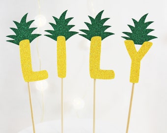 May include: A cake topper spelling out the name "LILY" with each letter decorated with a green glitter pineapple design.