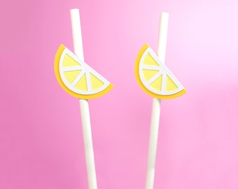 May include: Two white paper straws with yellow lemon slice cutouts attached to the top. The straws are against a pink background.