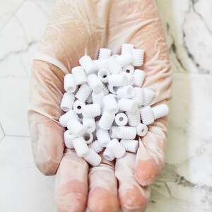 May include: A handful of small, white, cylindrical plastic components with ridged exteriors. The components are held in a gloved hand, with a bright, clean background.