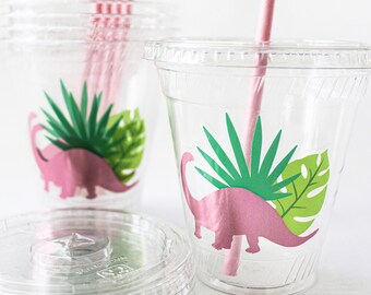 May include: Clear plastic cups with lids, decorated with pink dinosaur silhouettes and green tropical leaves. Pink paper straws are inserted in the cups. A stack of clear lids is in the foreground. Perfect for a dinosaur-themed party.