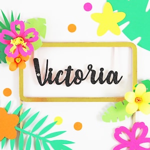 May include: A decorative cake topper with the name "Victoria" in black script, set within a gold frame. The design includes colorful paper flowers, green leaves, and confetti in shades of pink, yellow, and orange, creating a tropical theme.