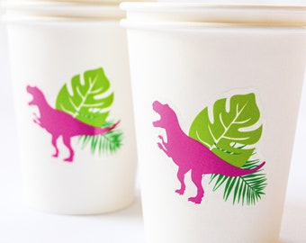 May include: White paper cups with a magenta Tyrannosaurus Rex graphic and green tropical leaves. The cups are designed for a party or event with a dinosaur theme. The cups are stacked and ready to be used.