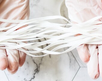 May include: A bundle of white elastic bands held by gloved hands. The bands are thin and flat, and they are arranged in a loose, flowing pattern. The background is a white and gray marble surface.