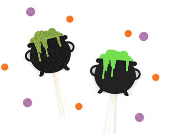 May include: Two black cauldron cupcake toppers with green slime on top. The toppers are on white sticks and are perfect for Halloween parties.
