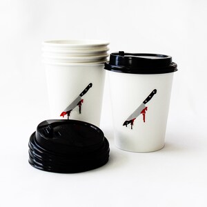 May include: Two white disposable coffee cups with black lids. One cup has a graphic of a bloody knife. The other cup is stacked on top of two other cups.