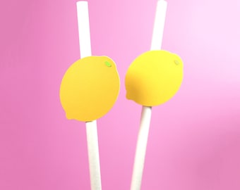 May include: Two white paper straws with yellow lemon cutouts attached to the top. The lemons have green centers. The straws are against a pink background.
