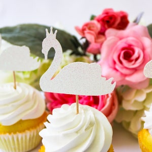 May include: White cupcakes with white frosting topped with glittery white swan cake toppers. The cupcakes are surrounded by pink and white artificial flowers.