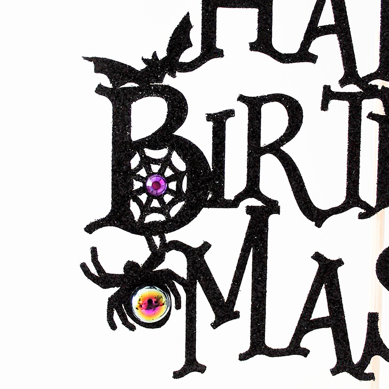 May include: Black glitter paper cut-out of the words "Happy Birthday" with a spider and spider web design. The spider has a purple and iridescent gem for an eye.