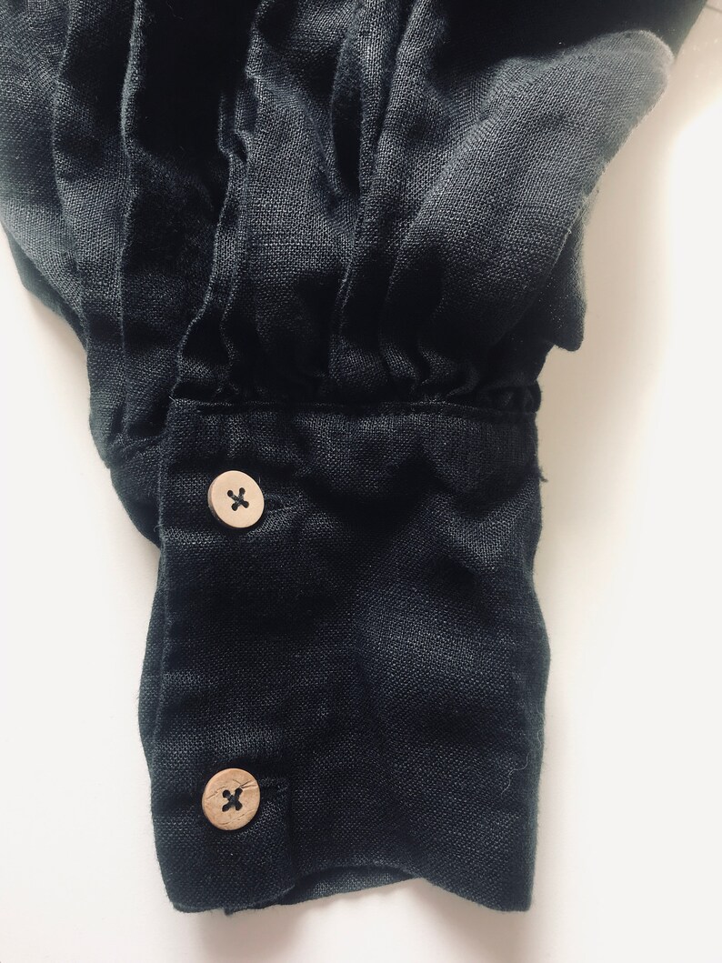May include: Close-up of a black linen shirt cuff with two wooden buttons. Each button has an 'X' etched into the surface.