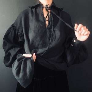 May include: A black linen shirt with a gathered neckline and long sleeves. The shirt has a tie at the neck and is tied in a knot at the front.