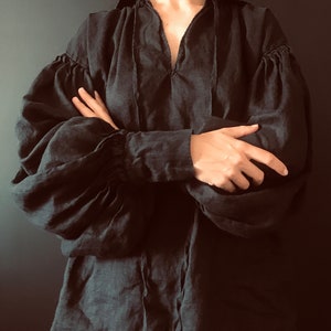May include: A black linen blouse with a gathered neckline and long, puffed sleeves. The blouse has a tie at the neckline and is cinched at the wrists.
