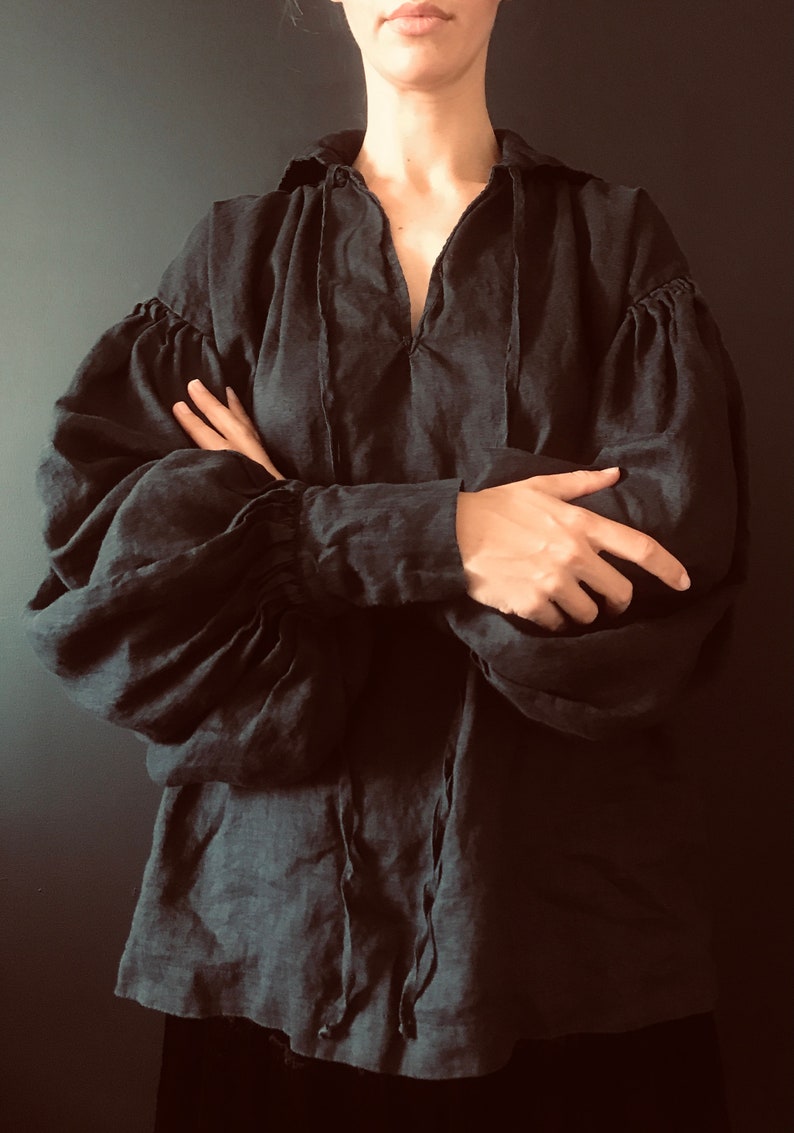 May include: A black linen blouse with a gathered neckline and long, puffed sleeves. The blouse has a tie at the neckline and is cinched at the wrists.