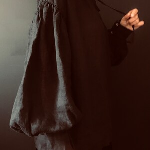 May include: A black linen shirt with a large, gathered puff sleeve. The shirt has a high collar and a tie at the neck.