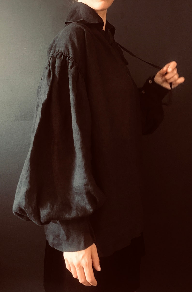 May include: A black linen shirt with a large, gathered puff sleeve. The shirt has a high collar and a tie at the neck.