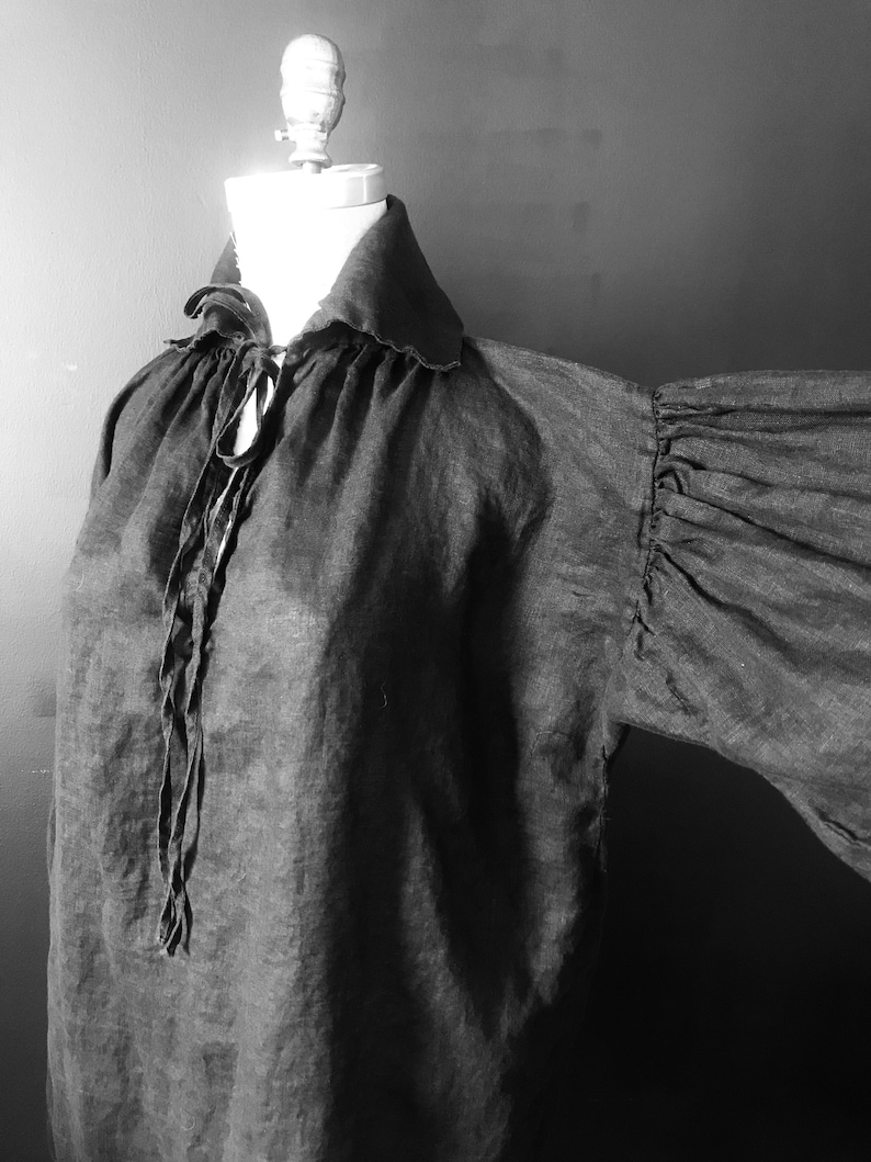 May include: A black linen shirt with a large, gathered collar and long, billowing sleeves. The shirt is tied at the neck with a long, thin cord.