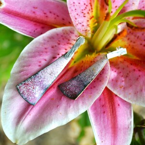 Puede incluir: Un par de pendientes de plata con un diseño moteado rosa y blanco, descansando sobre una flor de lirio rosa.