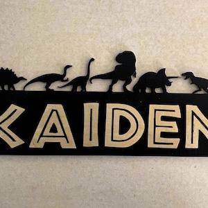 Könnte beinhalten: Ein schwarzes Holzschild mit dem Namen "KAIDEN" in goldenen Buchstaben. Das Schild ist mit Dinosauriersilhouetten und Logos für Jurassic Park und Jurassic World verziert.