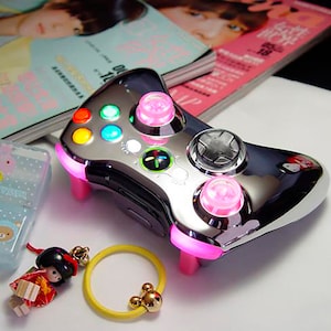 Puede incluir: Un mando de Xbox 360 cromado con botones rosas y una placa frontal de plástico transparente. El mando está sobre una superficie blanca.