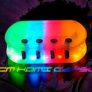 May include: A multi-colored, clear plastic power strip with three outlets. The power strip is illuminated with green, blue, and orange lights. The text "XCM HOMIC CAPSULE" is printed below the power strip.