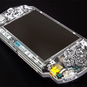 May include: A clear, transparent PlayStation Portable (PSP) handheld gaming console with visible internal components. The console has a black screen and a silver trim around the edges.