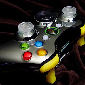 Puede incluir: Un mando Xbox 360 plateado y negro con detalles amarillos. El mando tiene joysticks de plástico transparente y botones de colores.