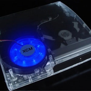 Puede incluir: Una carcasa transparente de plástico para una consola PlayStation 3 con un ventilador LED azul que dice "XCM".