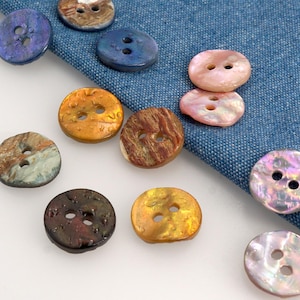 May include: A selection of decorative buttons in various colours and textures. The buttons are round with two holes and feature iridescent, marbled, and pearlescent finishes. Colours include blue, brown, gold, pink, and purple. The buttons are arranged on a blue fabric.
