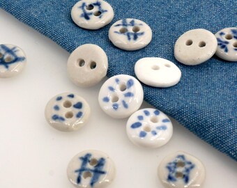 May include: A collection of ceramic buttons in various designs and colors. The buttons are primarily white with blue decorative patterns. Some buttons have two holes, while others have four. The buttons are scattered on a blue fabric surface.