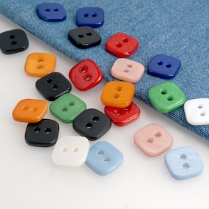 May include: A collection of square-shaped buttons in various colors, including blue, black, red, orange, green, pink, and white. Each button has two holes for sewing. The buttons are scattered on a blue fabric background.
