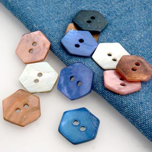 Op de afbeelding: Een verzameling iriserende, zeshoekige knopen in verschillende kleuren, waaronder blauw, roze, wit en bruin. Elke knoop heeft twee gaten om op te naaien en wordt gepresenteerd op een blauwe denim stoffen achtergrond.