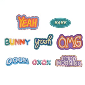 Op de afbeelding: Negen kleurrijke strijklabels met verschillende zinnen, waaronder "Yeah", "Babe", "Bunny", "Yoah", "OMG", "Oook!", "Oxox" en "Good Morning".