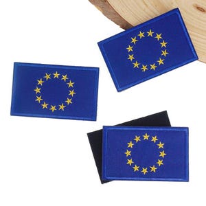 Pode incluir: Três patches retangulares azuis com a bandeira da União Europeia. Cada patch tem um círculo de doze estrelas amarelas sobre um fundo azul. Os patches são feitos de tecido e provavelmente para passar a ferro ou costurar.