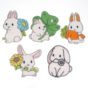 Op de afbeelding: Vijf geborduurde strijkpatches met schattige witte konijntjes met verschillende accessoires. De konijntjes houden een blauwe bloem, een groene klaver, een wortel, een zonnebloem en een witte bloem vast.