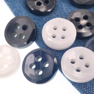 May include: Close-up of various buttons in shades of white, navy, and black. The buttons have either four holes or two holes. Some buttons have a faceted design, while others are round. The buttons are on a blue fabric.